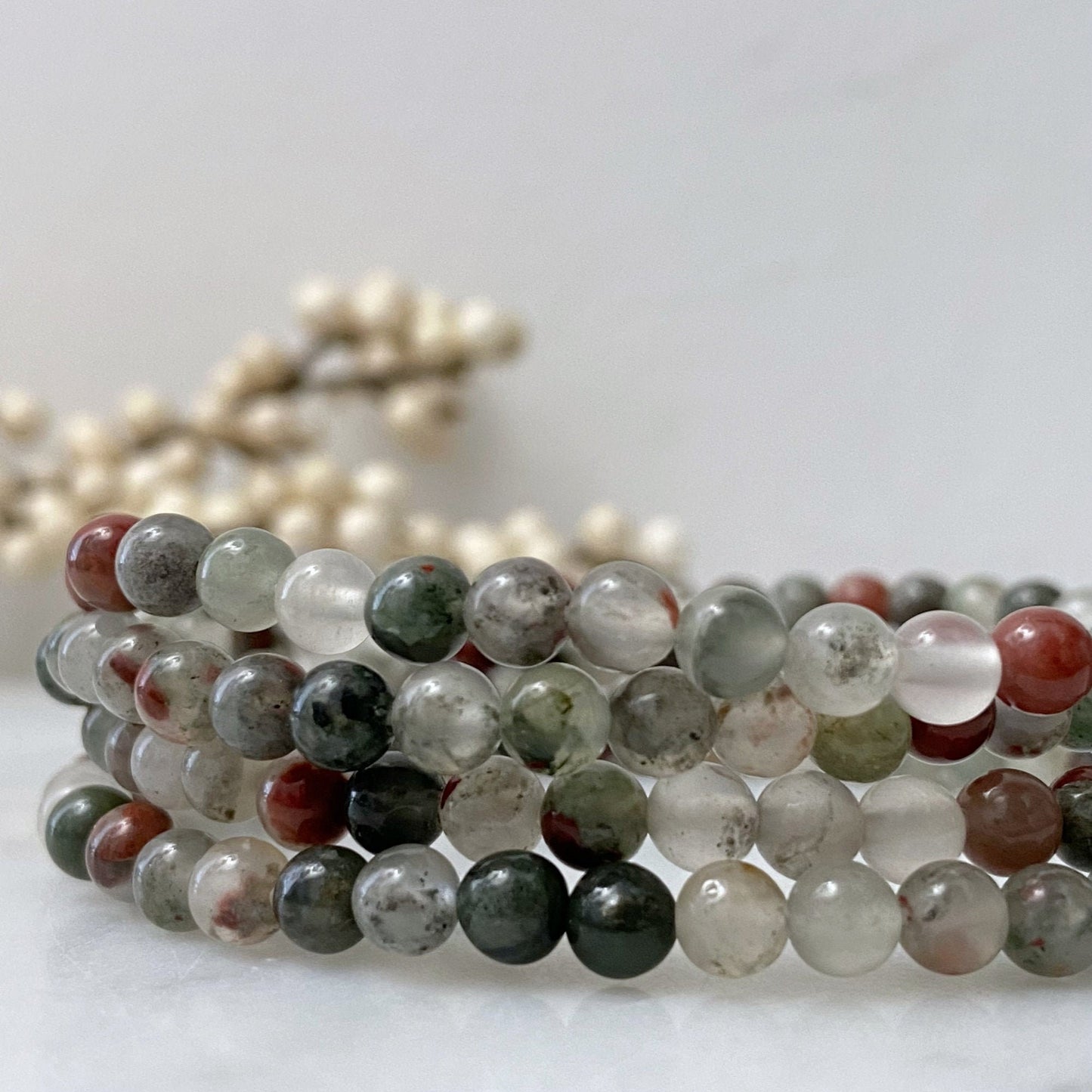 A close-up image of a stretchy, multi-strand 4mm Seftonite (African Bloodstone) Beaded Bracelet made of semi-transparent stones in various colors, including green, red, and grey. The bracelet is coiled neatly, exuding vibrant Reiki energy against a blurred backdrop featuring white, pearl-like beads.