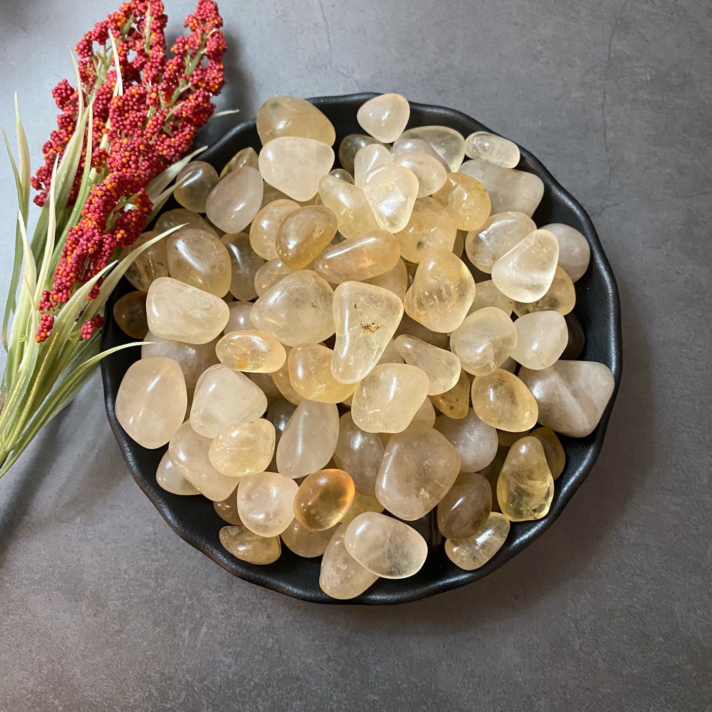 A black bowl filled with smooth, translucent yellow stones, possibly Natural Topaz Tumbled Stones known for their metaphysical properties, is placed on a gray surface. To the left of the bowl, a sprig of red and green artificial berries adds a pop of color to the scene.