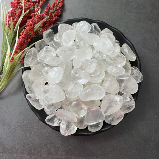 A black bowl filled with smooth, polished Clear Crystal Quartz Tumbled Stones is placed on a dark surface, perfect for crystal grids. To the left, there is a sprig of red and green dried flowers. The translucent stones emit a serene energy, ideal for healing rituals.