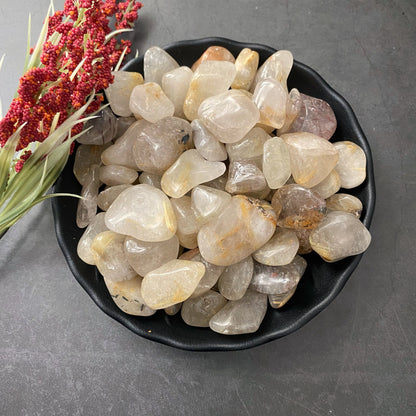 A black bowl filled with Rutilated Quartz Tumbled Stones of varying sizes, some showcasing subtle inclusions and color variations indicative of rutilated quartz. To the left, a small bouquet of red and green flowers rests on a dark, textured surface, enhancing the scene’s spiritual creativity.