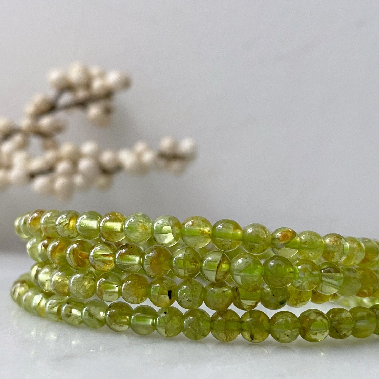 A close-up of a 4mm Peridot Beaded Bracelet made of translucent light green stones, coiled in multiple loops. The background is softly blurred, showcasing white berry-like ornaments, adding an elegant touch to the image while hinting at the bracelet's protective powers.