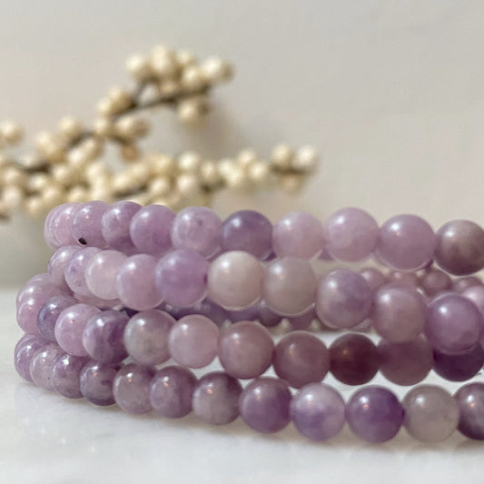 Close-up of a 4mm Lepidolite Beaded Bracelet made of polished, spherical purple stones. The bracelet is coiled in a circular shape, with a background featuring soft-focus white berry branches, adding a delicate and natural contrast to the image. Known for stress relief and chakra activation.