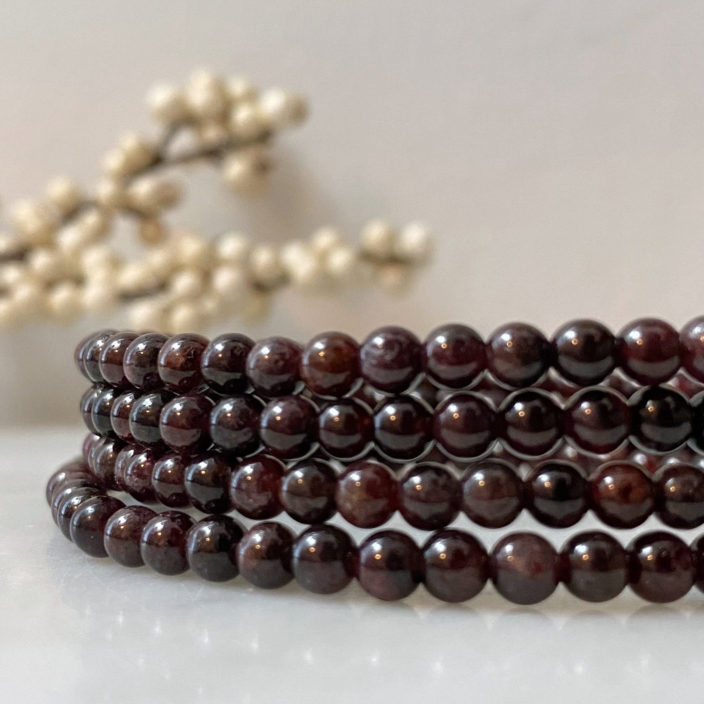 A stack of polished, 4mm Garnet Beaded Bracelets is displayed on a light surface. In the background, there is a blurred decorative element with white beads. The garnet beads are smooth and glossy, giving the bracelets an elegant appearance infused with Usui Reiki energy.