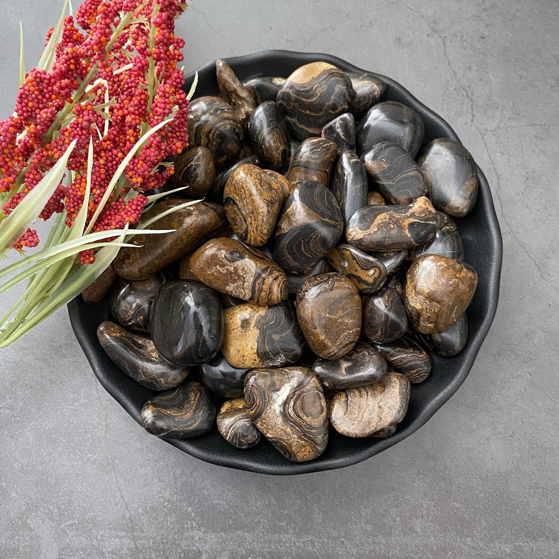 A black bowl filled with polished, brown and black Stromatolite Tumbled Stones sits on a gray surface. To the left, a bunch of red millet sprigs and green leaves adds a natural decorative touch. The scene sets a perfect ambiance for meditative sessions.