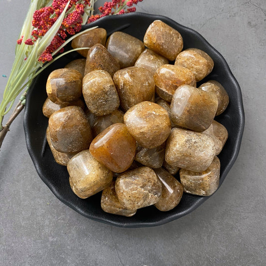 A black bowl brimming with polished Harlequin Quartz Tumbled Stones is placed on a gray surface. Red and white flowers rest beside the bowl, adding a touch of color to the arrangement and enhancing its emotional balancer properties.