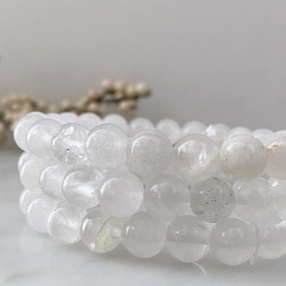 A close-up of a white beaded 8mm Clear Quartz Beaded Bracelet made of spherical semi-transparent stones, neatly coiled with blurred background details, which include out-of-focus beige elements. The clear quartz polished beads appear smooth and polished, with natural variations in clarity.