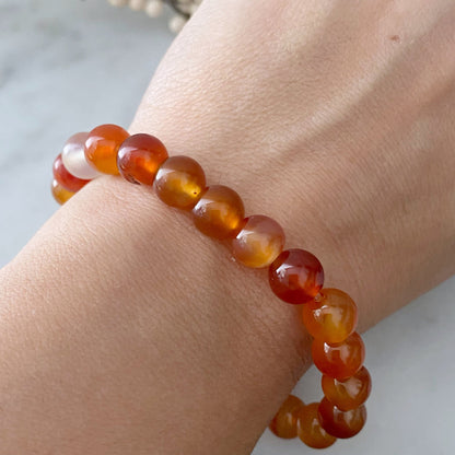 A round 8mm Carnelian Agate Beaded Bracelet made from translucent orange stones is placed on a reflective black surface, creating a mirrored effect. This fertility crystal is composed of smooth, polished beads, varying in shades of orange and yellow.