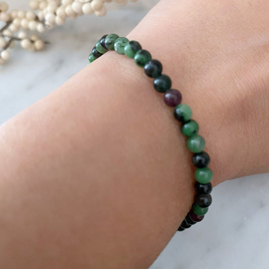 A close-up photo of a person wearing a 4mm Ruby Zoisite Beaded Bracelet on their wrist. The bracelet features polished round beads in varying shades of green, black, and a hint of purple. The background is slightly blurred with some out-of-focus decoration visible.