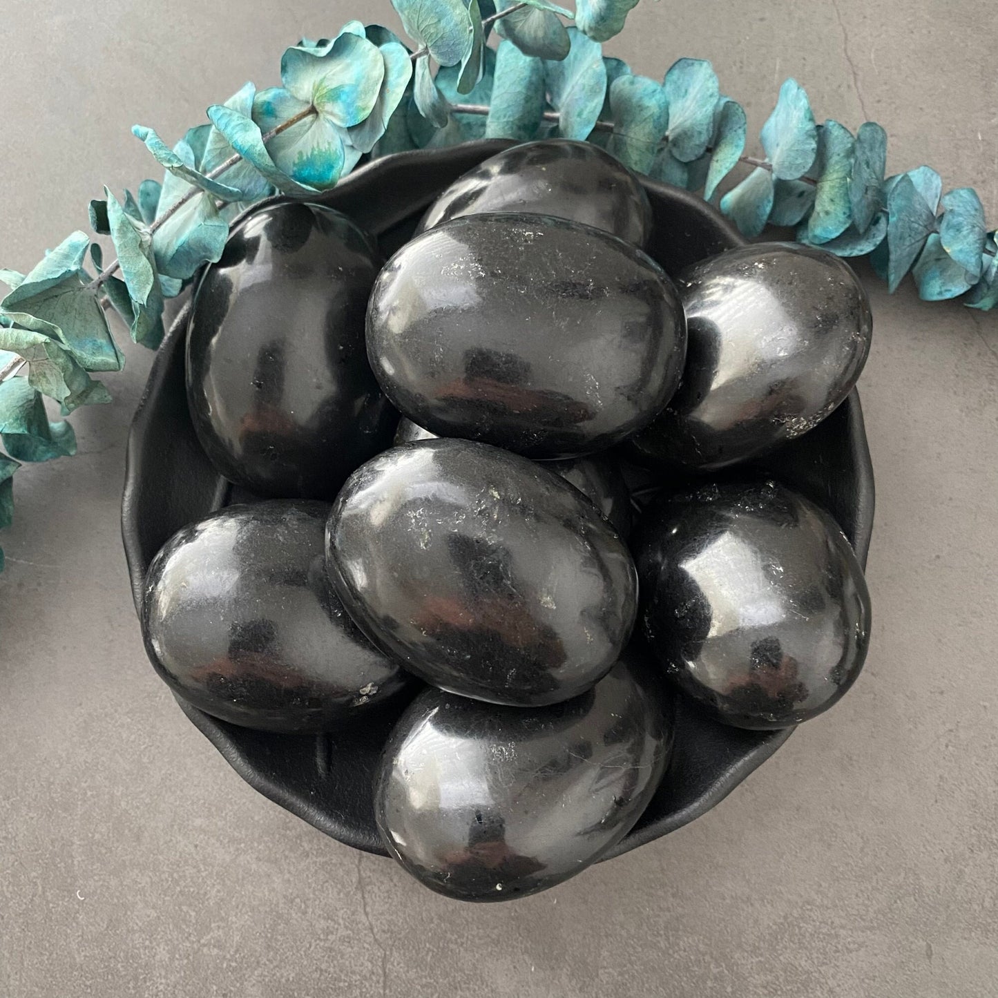 A black bowl filled with several shiny black eggs is placed on a gray surface. Blue-green leaves are arranged around the bowl, creating a decorative border. The eggs' polished surfaces reflect light, resembling Black Tourmaline Palm Stone and adding grounding properties to the composition.