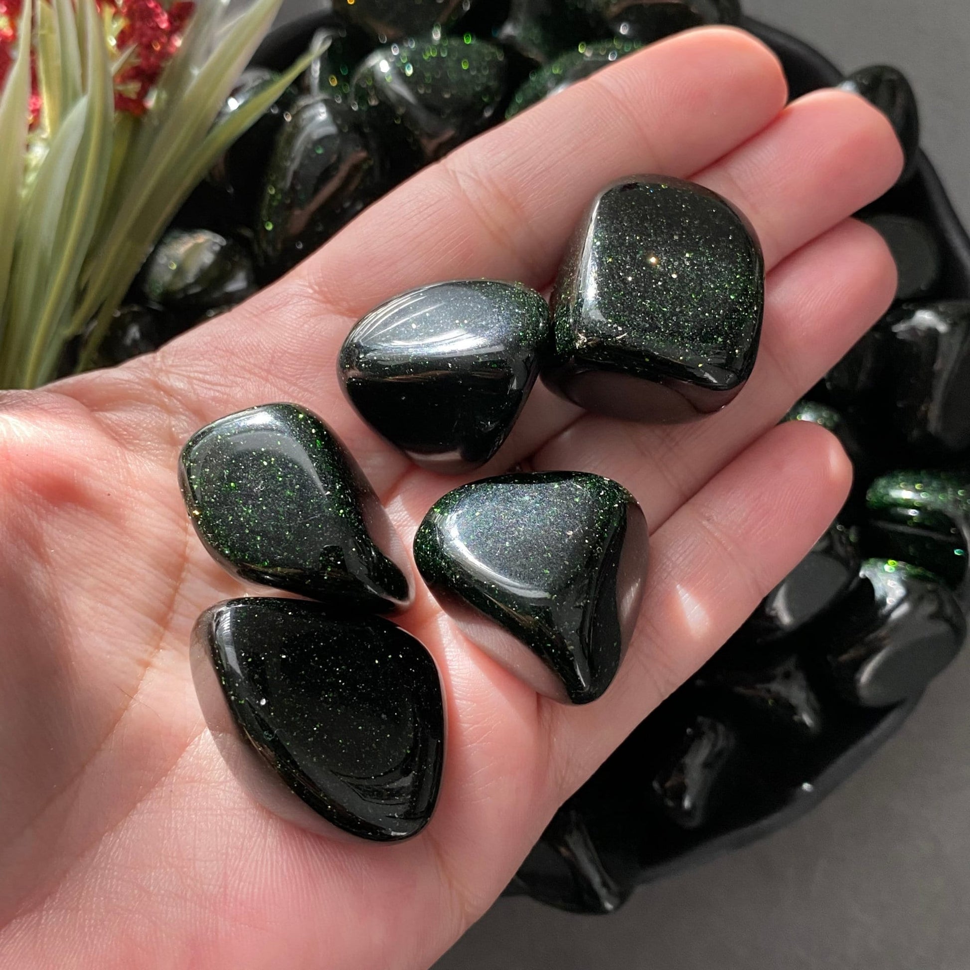 A black bowl filled with shiny, polished black stones sits on a gray surface. To the left of the bowl, a sprig of green leaves and red berries adds a touch of color to the composition. Among the stones are Green Goldstone Tumbled Stones, known as protection minerals that balance Heart Chakra crystals.