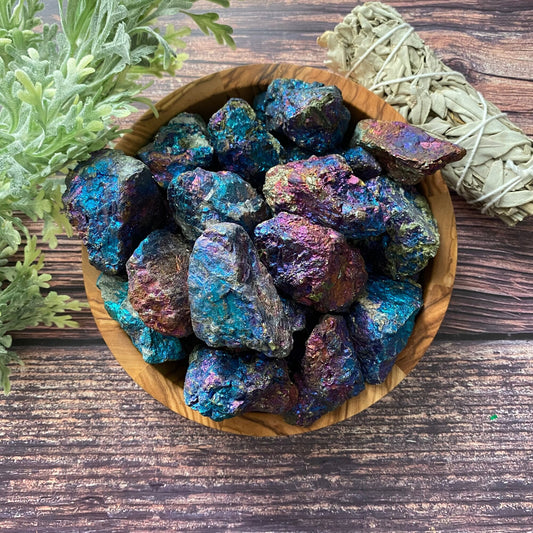 A wooden bowl filled with large, iridescent Raw Chalcopyrite Peacock Ore Stones, also known as Peacock Ore for their vibrant blue, purple, and green hues, sits on a rustic wooden surface. A bundle of sage and green foliage can be seen in the background.