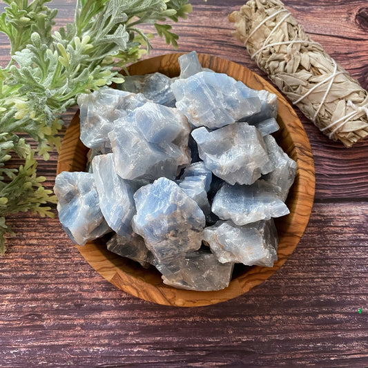 A wooden bowl filled with rough, light blue Raw Blue Calcite Stones is placed on a wooden surface. To the left of the bowl, there is a bunch of green leaves, and to the right, a bundle of sage wrapped in string is visible.