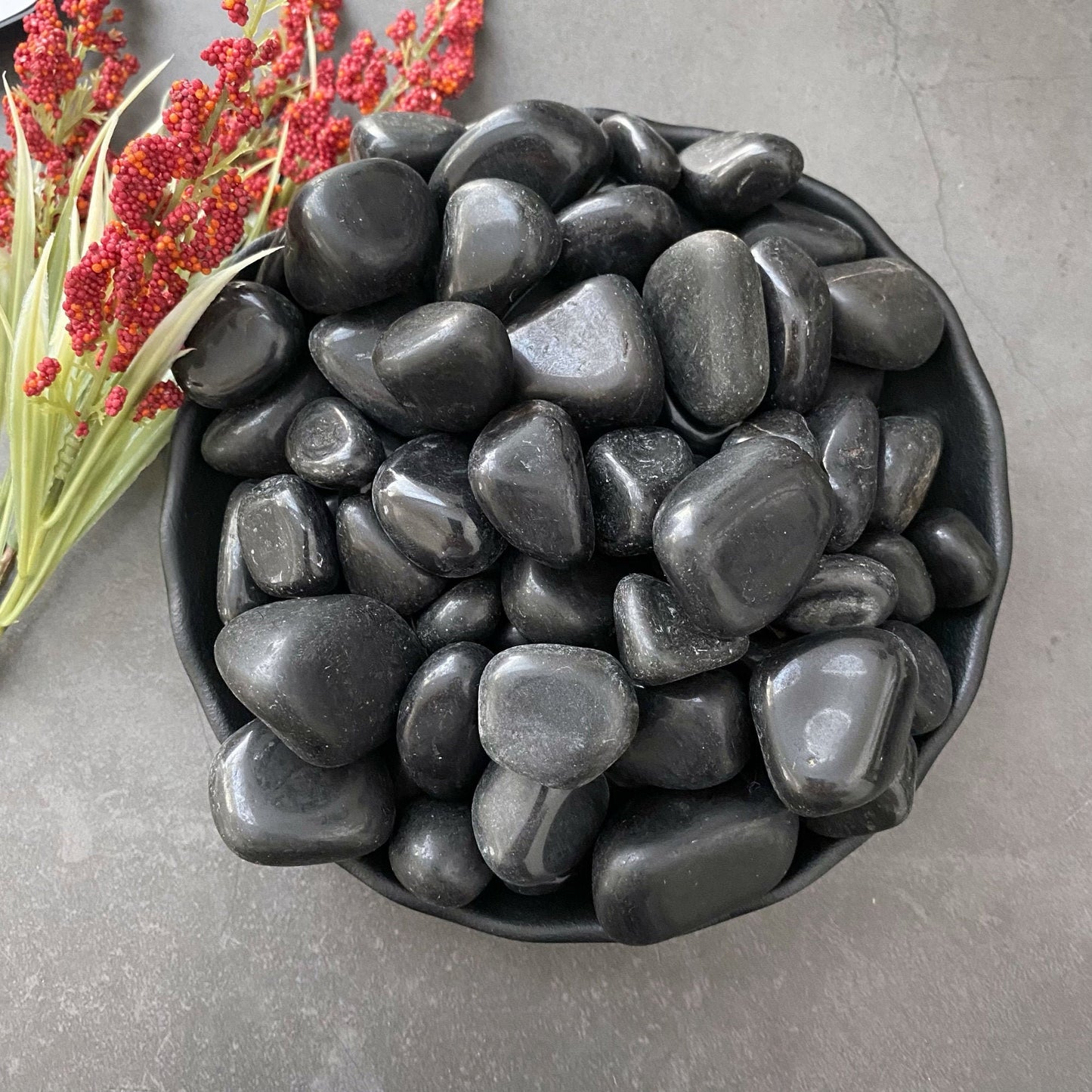 A black bowl filled with smooth, rounded Black Tourmaline Tumbled Stones is placed on a gray surface. Next to the bowl, there is a bunch of red and green dried flowers, adding a touch of color to the scene while enhancing its metaphysical properties.