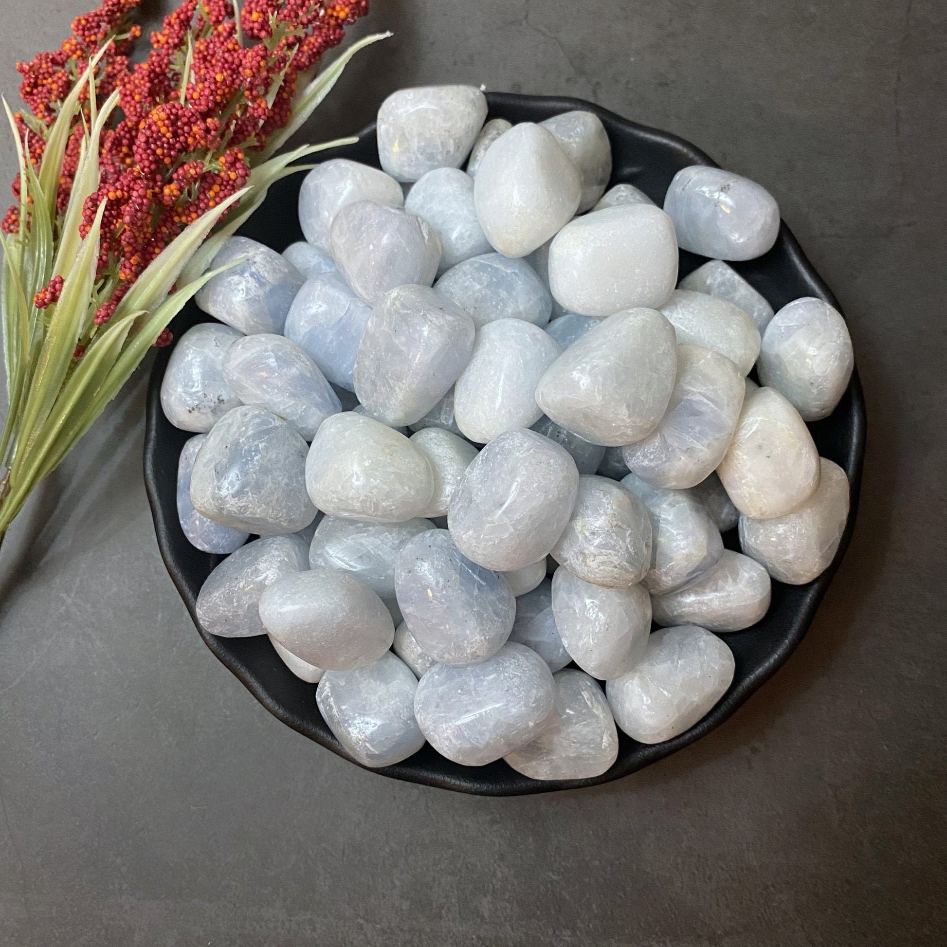 A black plate full of smooth, rounded Blue Calcite Tumbled Stones sits on a dark surface. Beside the plate is a bunch of red berries with green leaves. The arrangement, resonating with Throat Chakra energy, gives a natural and calming aesthetic.