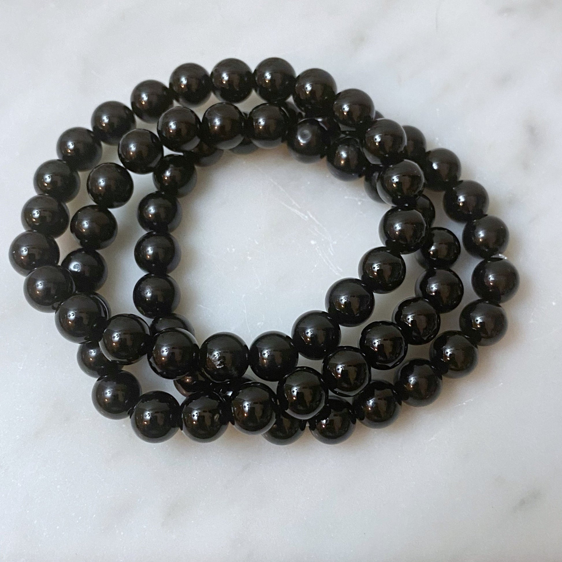 A beautiful 8mm Black Onyx Beaded Bracelet made of glossy black beads is displayed on a white surface. In the background, out of focus, there's a delicate branch with small white berries. The composition creates a pleasing contrast between the shiny beads and the soft white berries, emanating psychic protection.