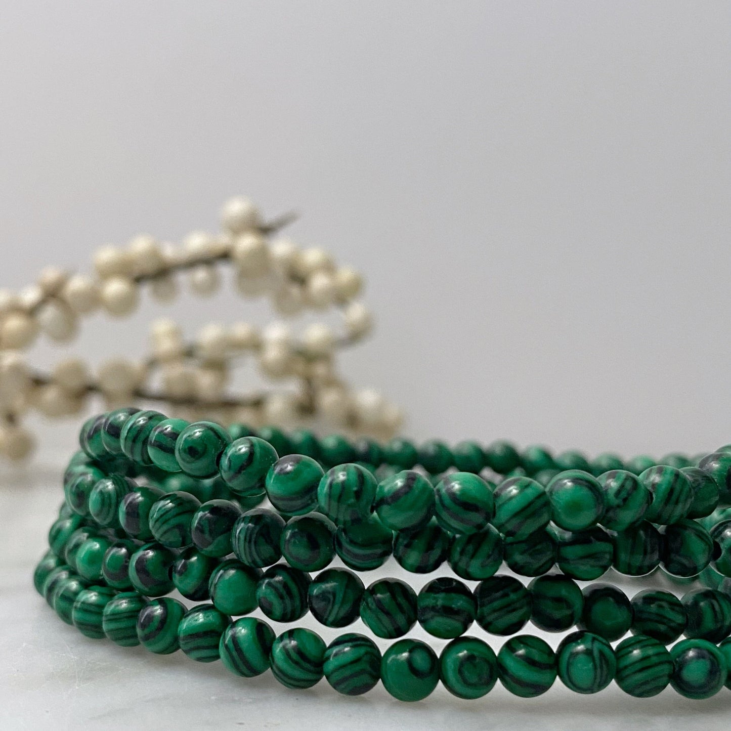 A 4mm Malachite Beaded Bracelet is stacked neatly on a smooth surface. The background is minimal with a blurred, light-colored beaded decoration adding a soft touch to the composition.