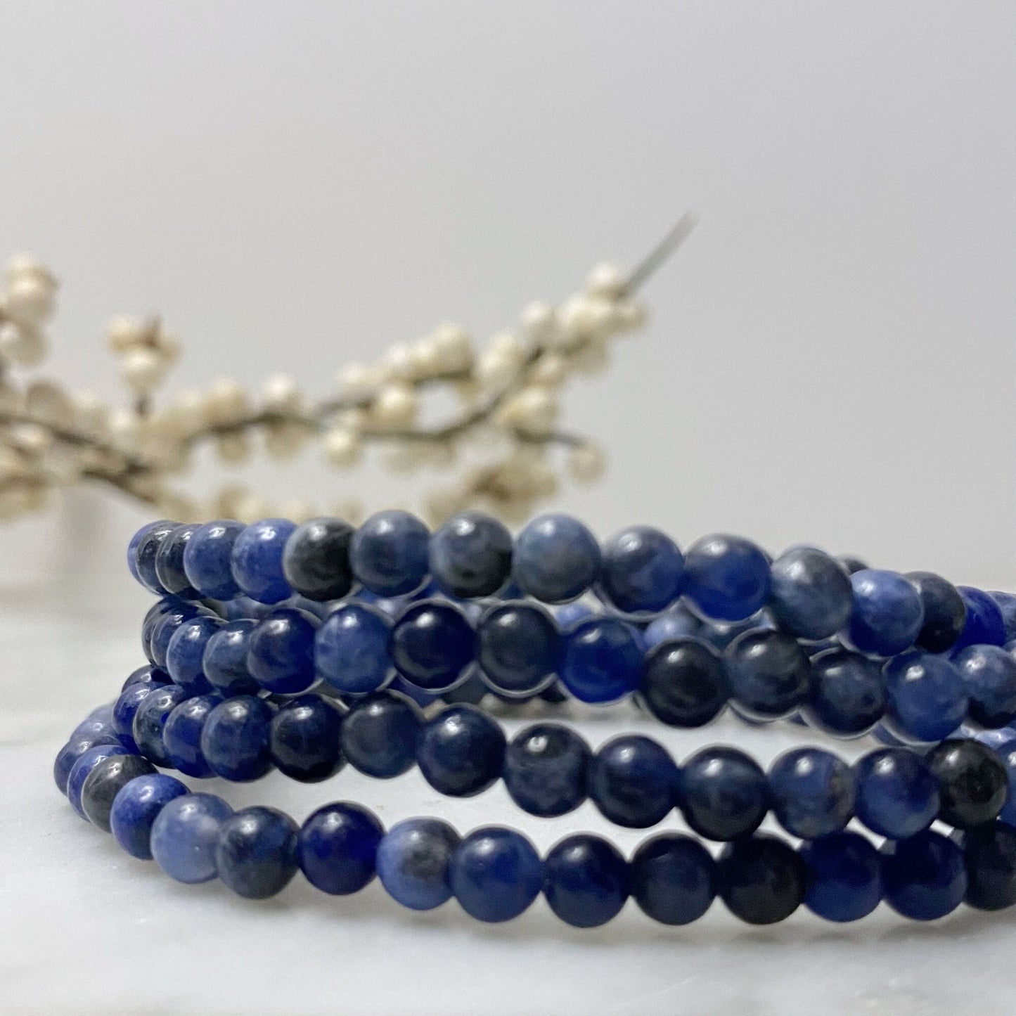 A stack of 4mm Sodalite Beaded Bracelets is displayed on a white marble surface. The deep blue beads have slight variations in color, adding texture to the bands. In the blurred background, delicate white buds on branches add a soft contrast, promoting emotional balance and healing Usui Reiki energy.