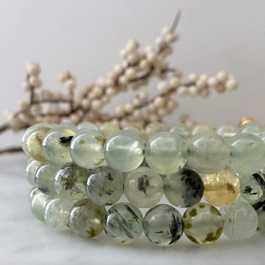 A close-up shot of an 8mm Prehnite Beaded Bracelet made from translucent green beads with dark inclusions. The bracelet is coiled in multiple loops on a light grey surface. In the background, a blurred branch with small, off-white buds is visible, creating a soft, natural ambiance.