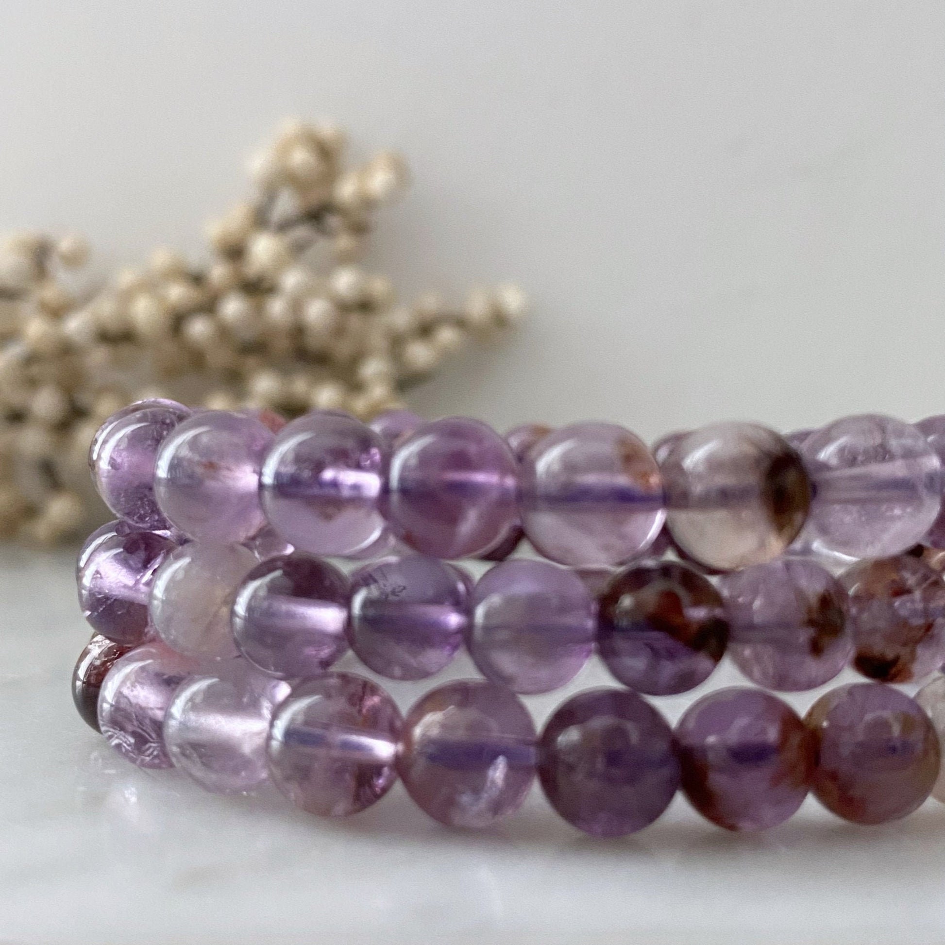 A circular 8mm Super Seven Beaded Bracelet displayed on a glossy black surface, with each bead featuring a mix of translucent purple and reddish hues. The bracelet's reflection is visible below on the polished surface, enhancing its vibrant colors.