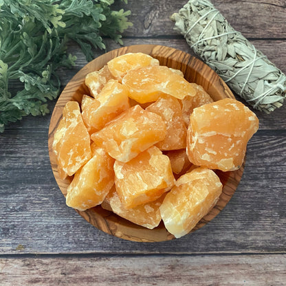 A wooden bowl filled with large, rough chunks of Raw Orange Calcite Stones sits on a wooden surface, radiating creative energies. To the right, there's a bundle of dried sage wrapped in twine. Green foliage is partially visible in the top left corner.