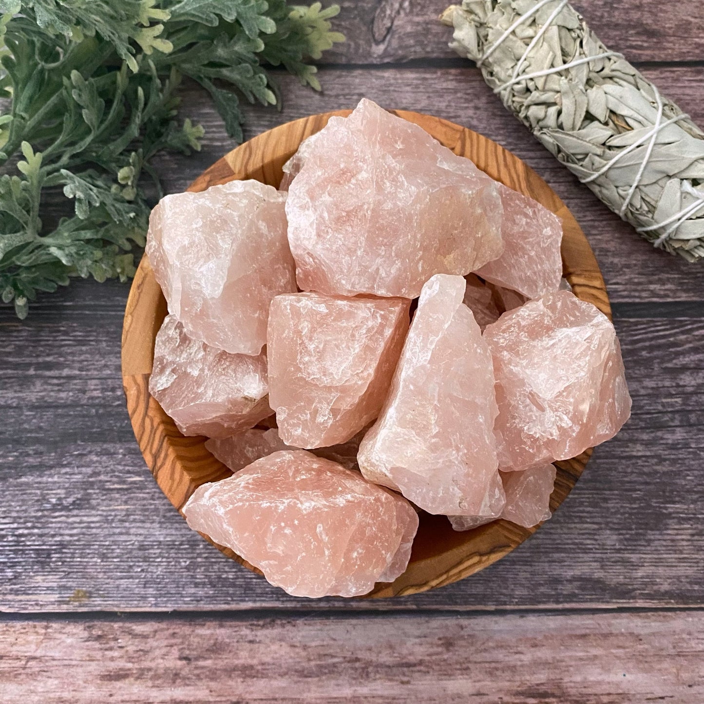 A wooden bowl filled with large chunks of Raw Rose Quartz Stones is placed on a wooden surface. To the left, green foliage is visible, and to the right, there is a bundle of sage for smudging, emphasizing the setting's focus on emotional healing and unconditional love.