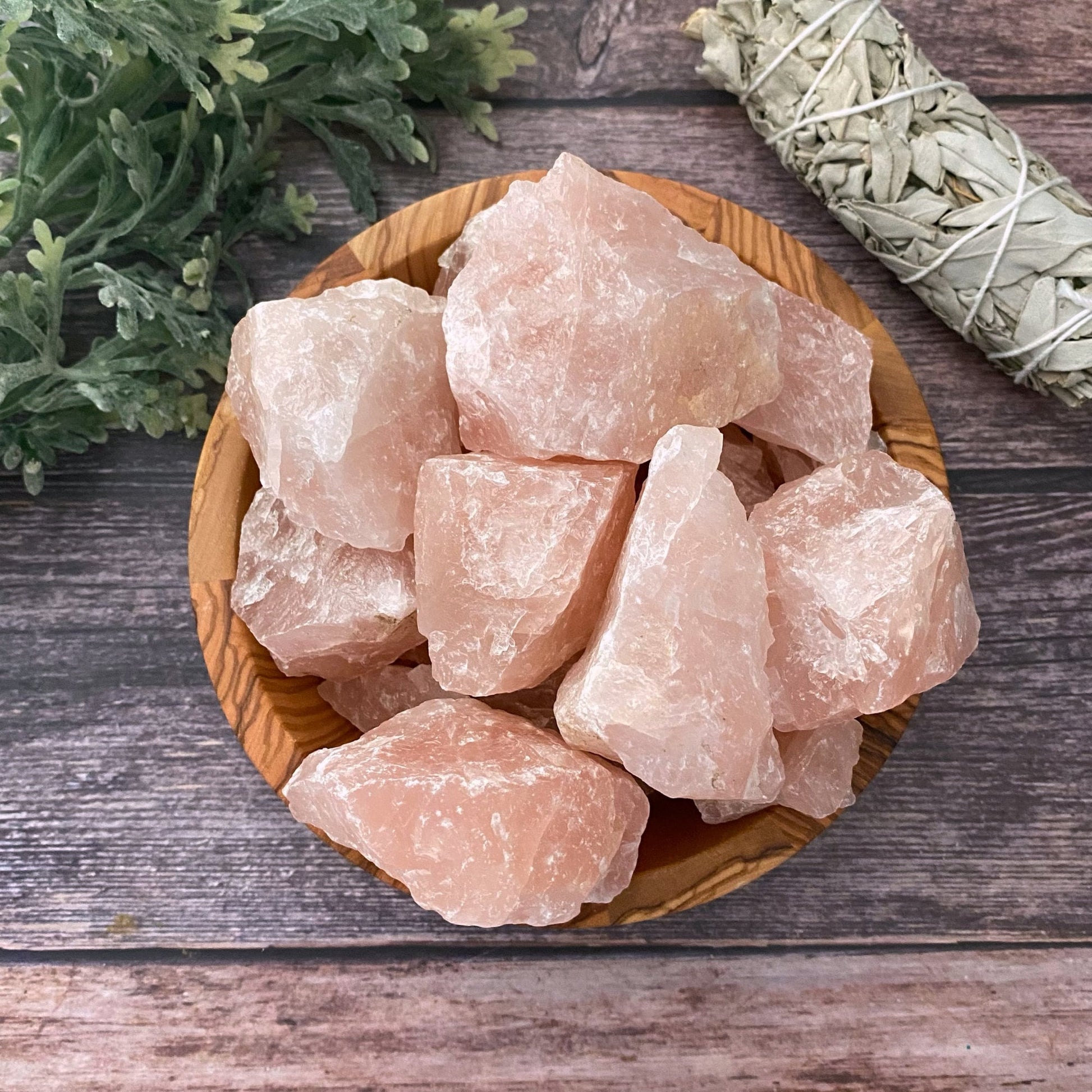 A wooden bowl filled with large chunks of Raw Rose Quartz Stones is placed on a wooden surface. To the left, green foliage is visible, and to the right, there is a bundle of sage for smudging, emphasizing the setting's focus on emotional healing and unconditional love.