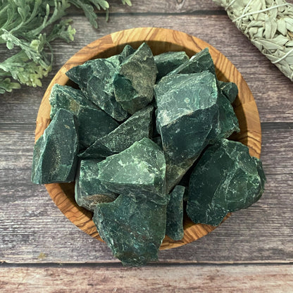 A wooden bowl holds an assortment of large, rough, dark green jade stones on a wooden surface. To the top left, green foliage is visible, and to the right, a bundle of dried sage rests. Among the jade stones lie Raw Bloodstone Stones prized for their spiritual applications and healing properties.