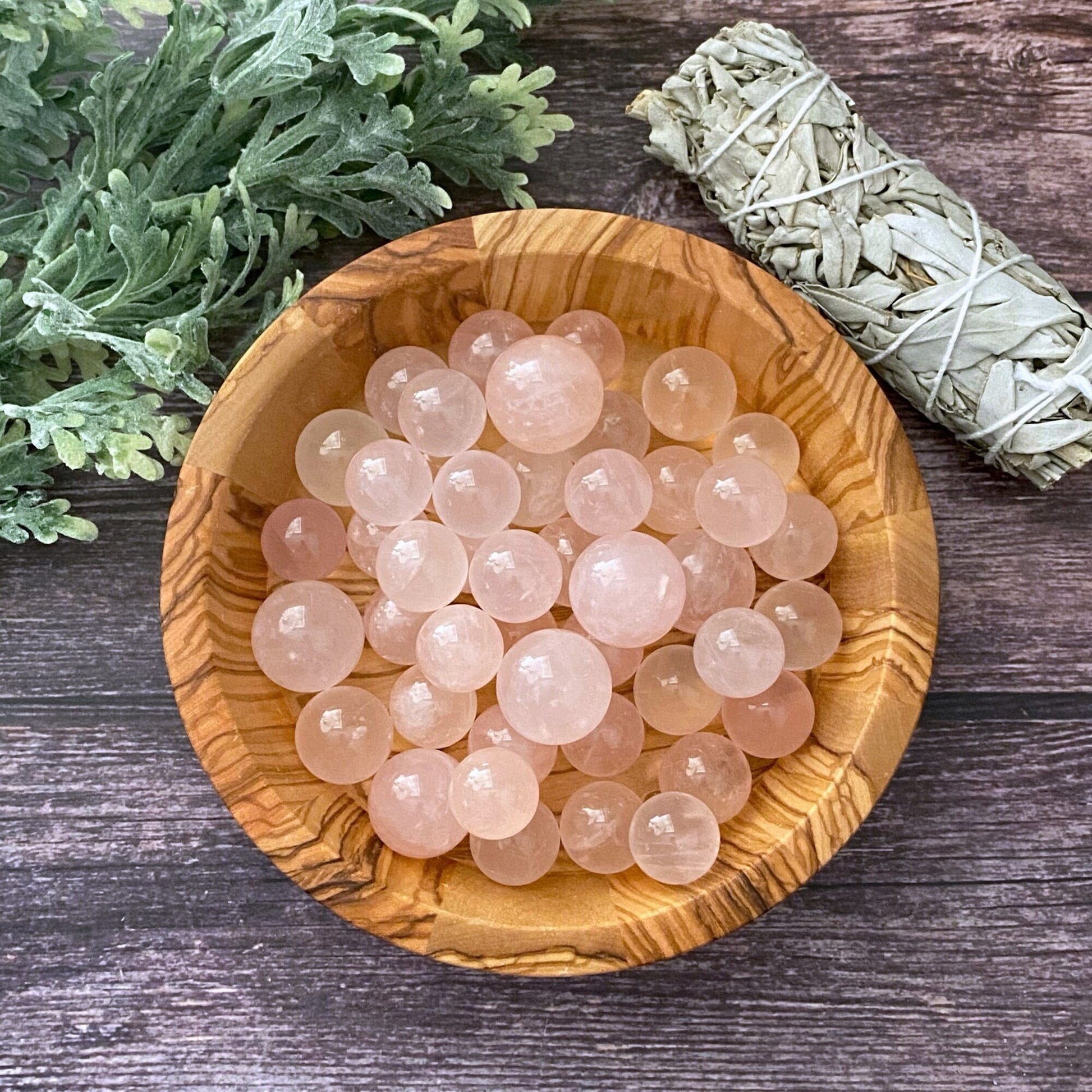 A wooden bowl filled with smooth, round Mini Rose Quartz Spheres is placed on a wooden surface with a bundle of sage and green leafy plant to the side. The bowl and its contents are centrally positioned, creating a harmonious and earthy display perfect for meditation.