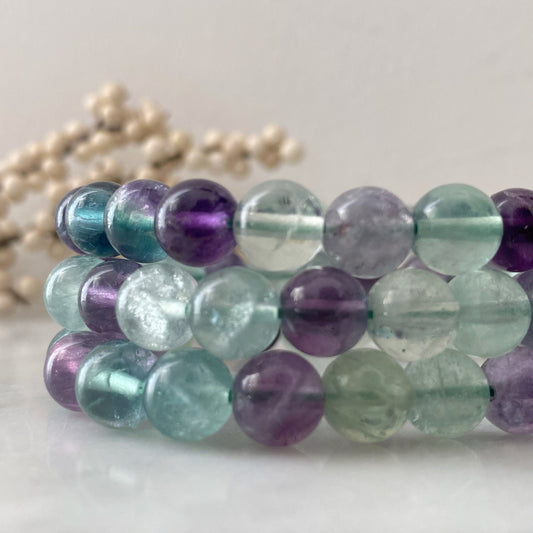 A close-up image of three stacked 8mm Fluorite Beaded Bracelets, crafted from polished beads in shades of purple, green, and blue. The background is blurred, with strands of white pearls faintly visible. The surface is white and glossy.