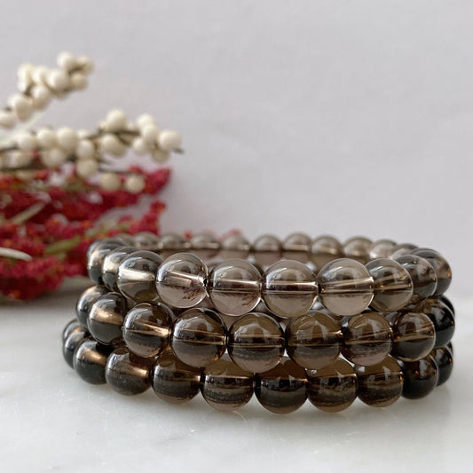 A close-up image shows several stacked 8mm Smoky Quartz Beaded Bracelets made of round, translucent beads. In the background, there are decorative elements, including small off-white and red dried flowers against a plain, light-colored surface. These grounding stone bracelets exude calming Usui Reiki energy.