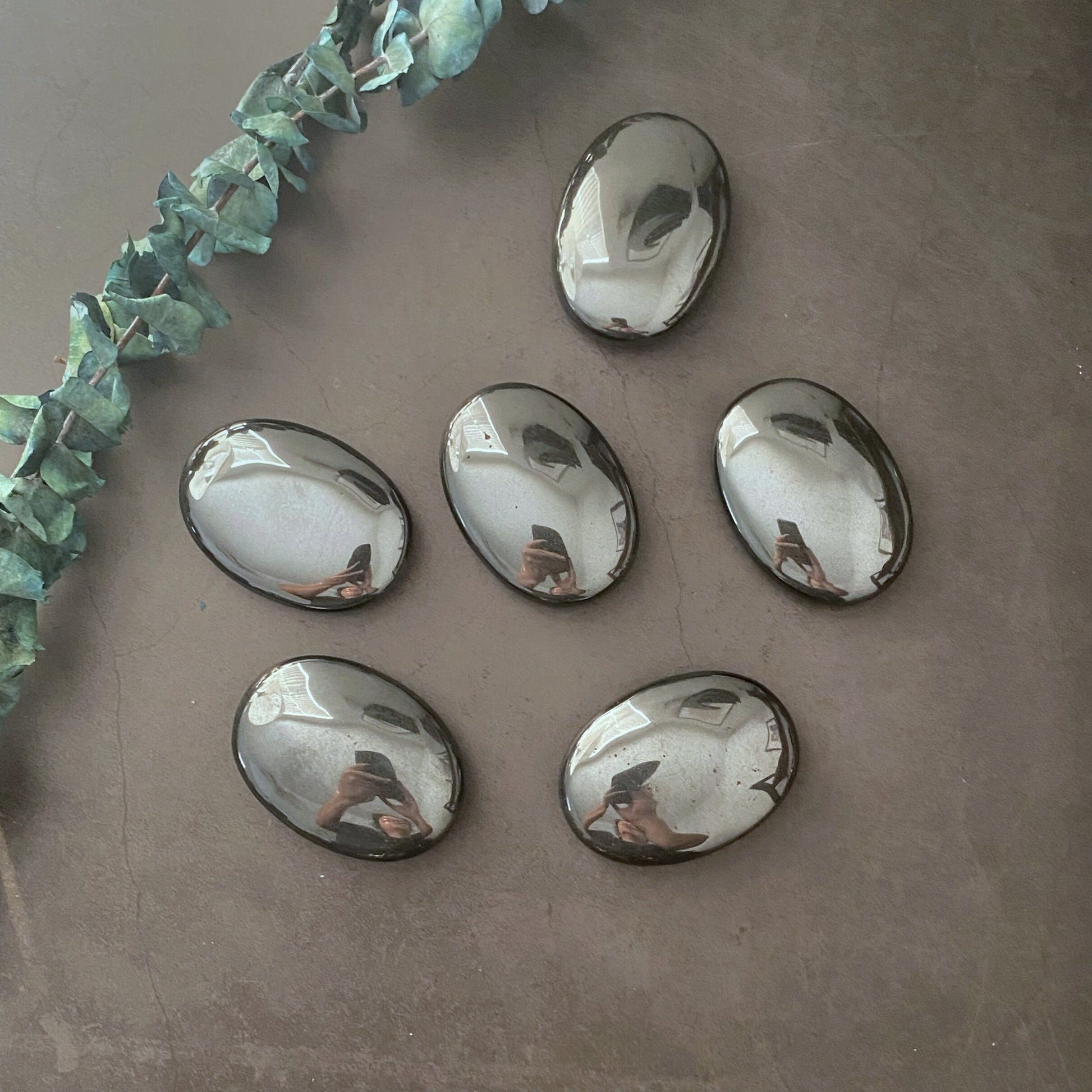 A black bowl containing five 2.75" Hematite Palm Stones with a reflective surface. The stones, known for their grounding properties, mirror the surrounding environment. A piece of green foliage rests beside the bowl on a dark, solid background.