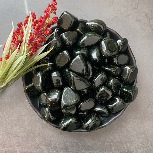 A black bowl filled with shiny, polished black stones sits on a gray surface. To the left of the bowl, a sprig of green leaves and red berries adds a touch of color to the composition. Among the stones are Green Goldstone Tumbled Stones, known as protection minerals that balance Heart Chakra crystals.