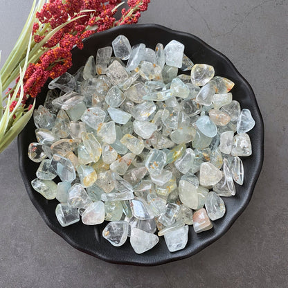 A black bowl on a gray surface is filled with Blue Topaz Tumbled Stones, clear and light-colored, possibly quartz. Some stones have slight yellow and green tints. Red and yellow sprigs lie beside the bowl.