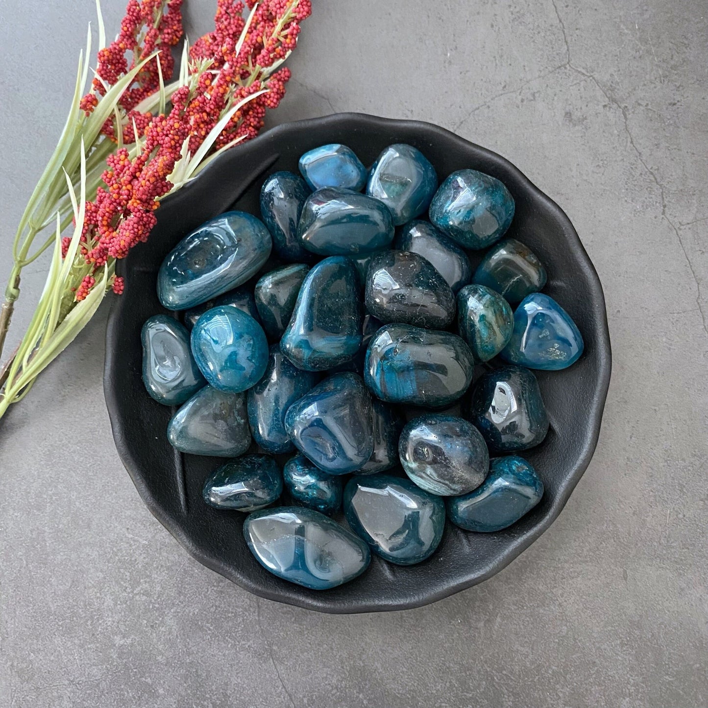 A black bowl filled with polished blue stones, ideal for balancing the throat chakra, rests on a grey textured surface. To the left of the bowl are sprigs of red berries with green stems. The shiny, smooth stones showcase various shades of blue, including Teal Agate Tumbled Stones, known as a calming stone.