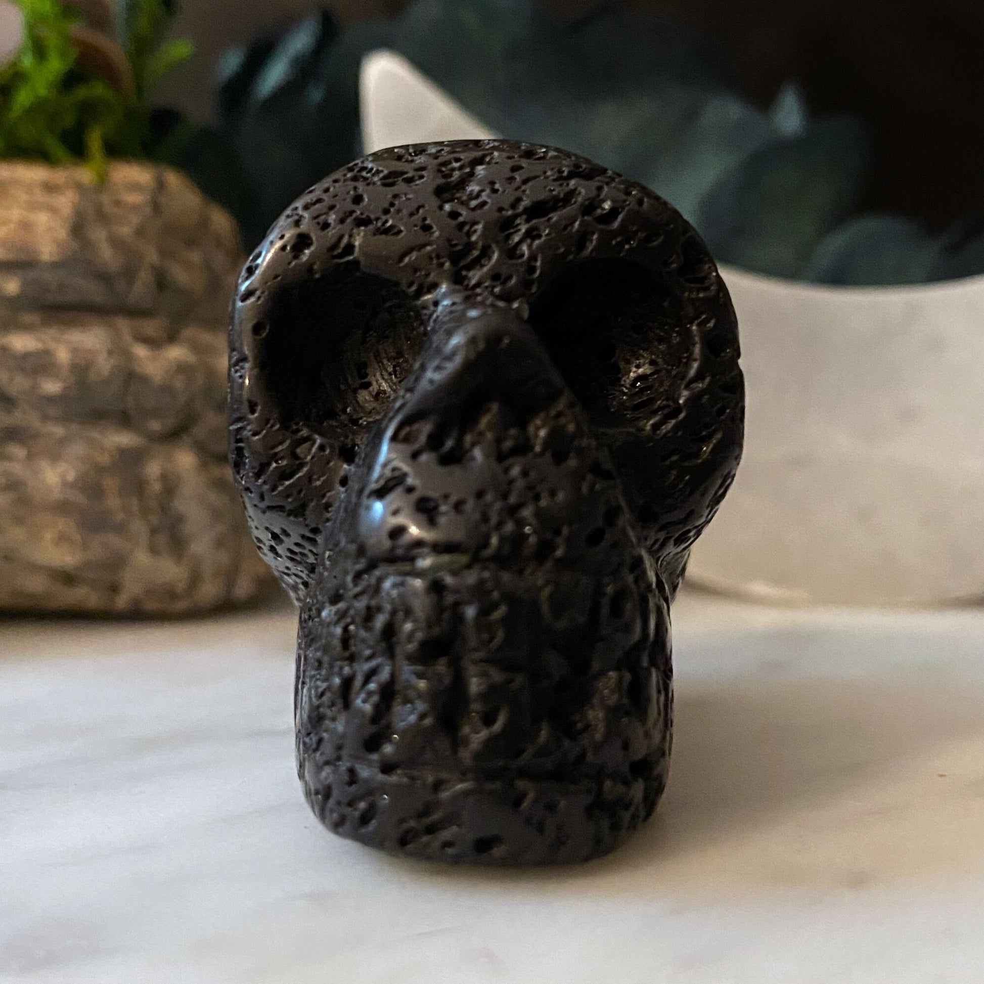 A 2" Lava Rock Skull is displayed on a flat surface with a blurred background of other natural stone formations and greenery. The texture of the skull is rough and dotted with numerous small holes, resonating with energies that ground and activate the root chakra.