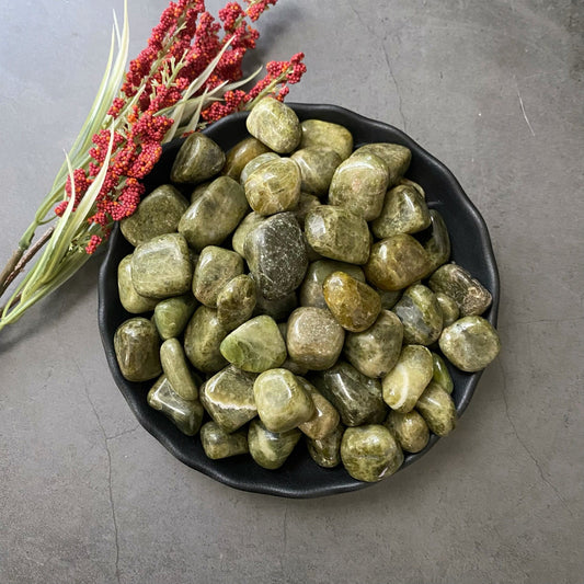 A black bowl filled with smooth Vesuvianite Tumbled Stones sits on a gray surface. Next to the bowl, there is a sprig of red berries with green leaves, adding a touch of color to the scene. These stones are known for promoting Heart Chakra balance and emotional healing.