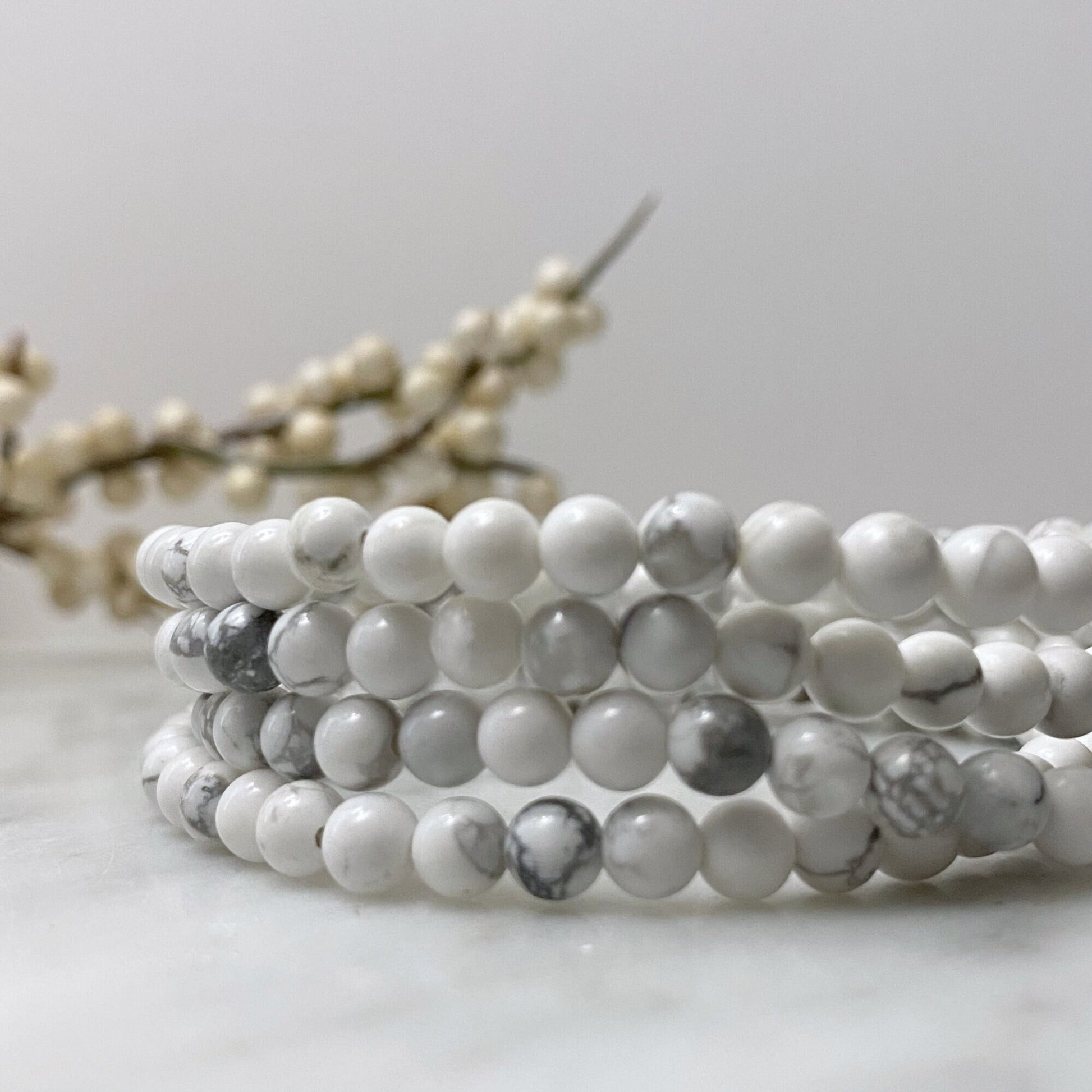 A 4mm Howlite Beaded Bracelet made of white marble-like stones with subtle gray veining is displayed against a black reflective surface, highlighting the bracelet's elegant and smooth texture. It’s perfect for those seeking emotional healing and balance through Usui Reiki energy.