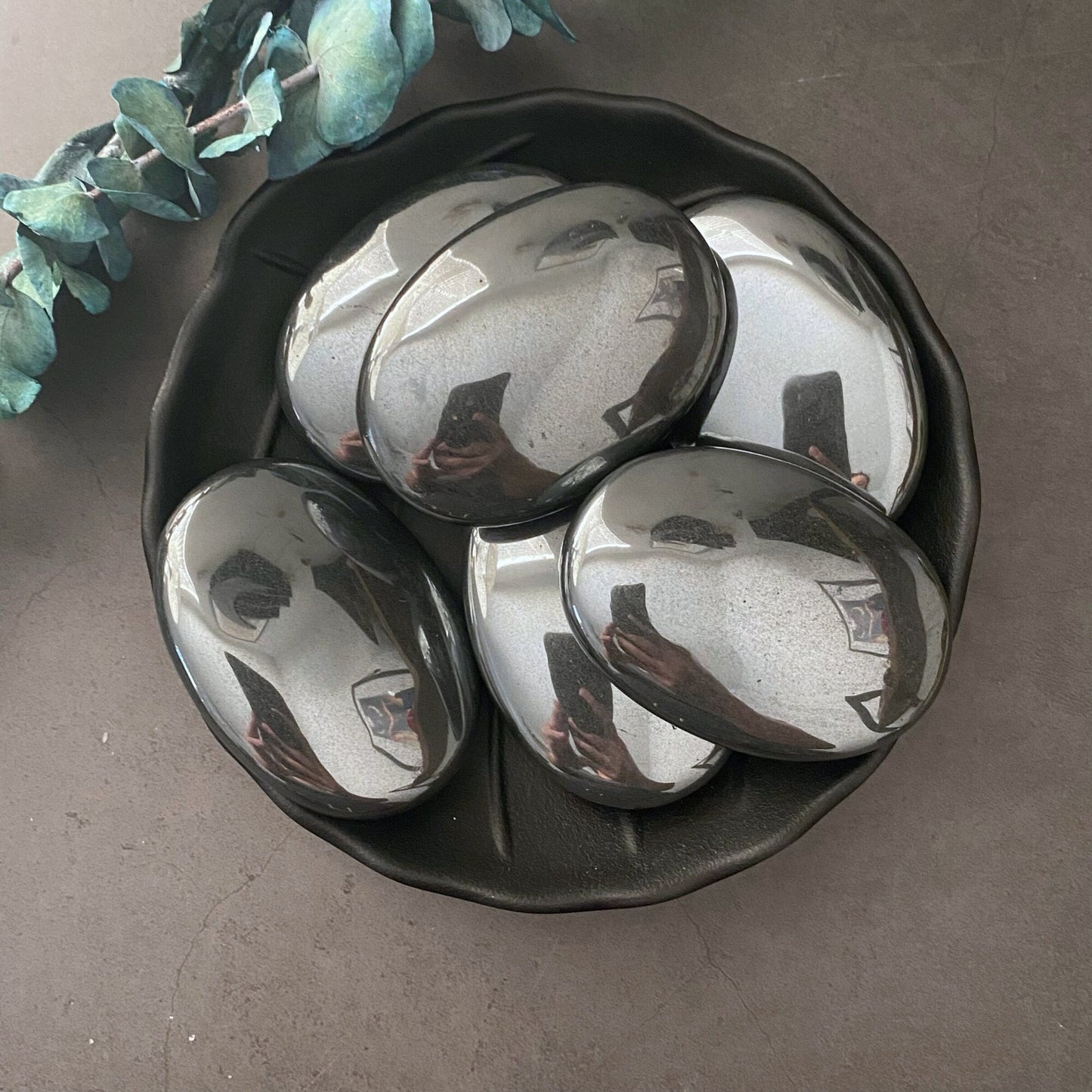 A black bowl containing five 2.75" Hematite Palm Stones with a reflective surface. The stones, known for their grounding properties, mirror the surrounding environment. A piece of green foliage rests beside the bowl on a dark, solid background.