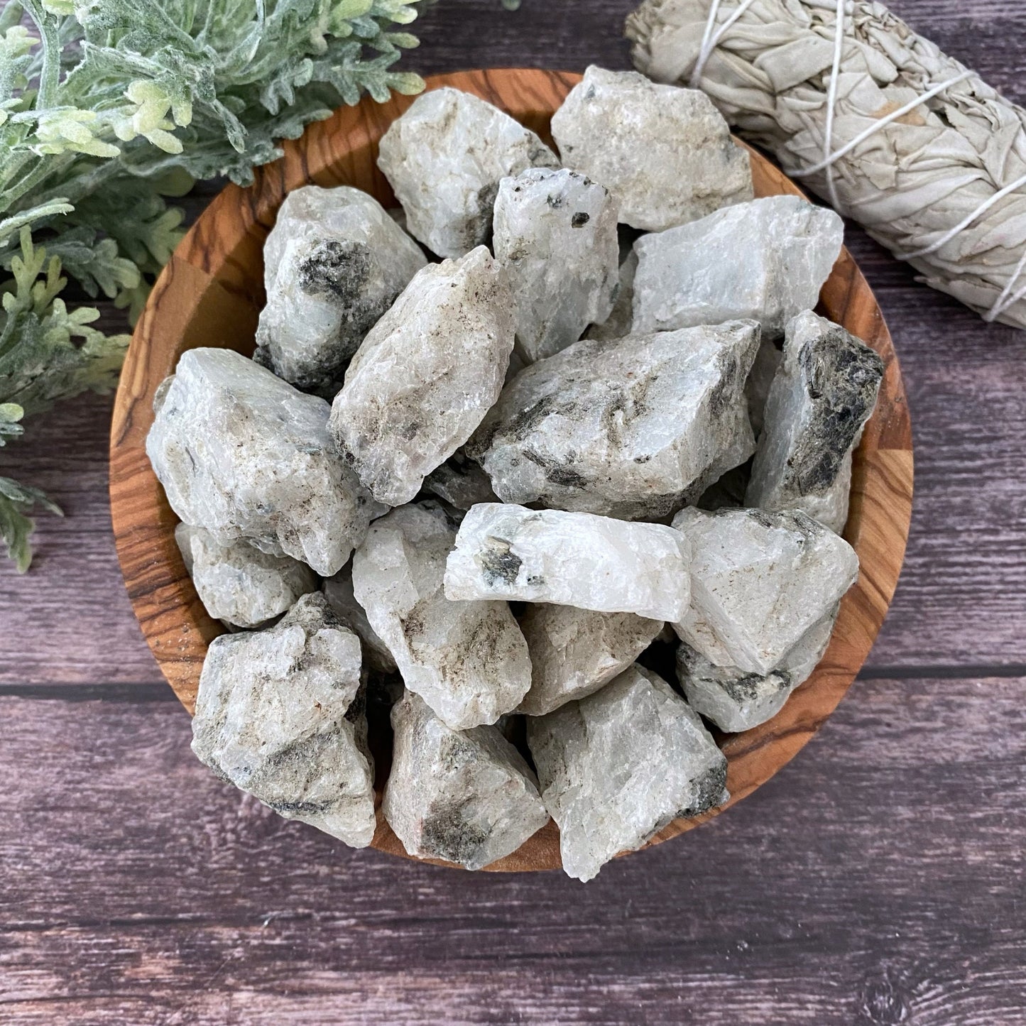 A wooden bowl filled with Raw Rainbow Moonstone Stones is placed on a wooden surface. Sprigs of greenery and a bundle of dried sage are in the background, adding a natural and earthy feel to the scene, perfect for enhancing psychic protection.