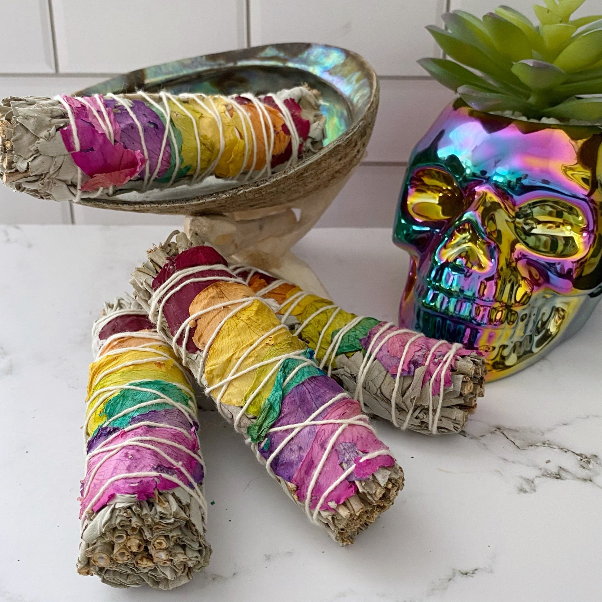 Three bundles of Organic White Sage & Chakra Rose Petals wrapped with colorful thread are arranged in front of a decorative metallic skull and a small potted succulent. Behind the sage bundles, there is a shell on a stand. The background features white tile, creating an ideal setting for energy cleansing with these chakra smudge sticks.