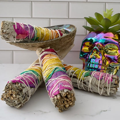 Three bundles of Organic White Sage & Chakra Rose Petals wrapped with colorful thread are arranged in front of a decorative metallic skull and a small potted succulent. Behind the sage bundles, there is a shell on a stand. The background features white tile, creating an ideal setting for energy cleansing with these chakra smudge sticks.