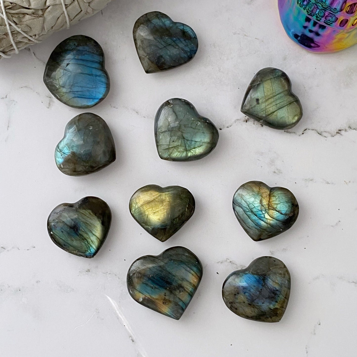 A collection of 1.5" Small Labradorite Hearts with Flash is arranged on a white marble surface. In the background, a bundle of sage and a multicolored, reflective skull decoration are visible.