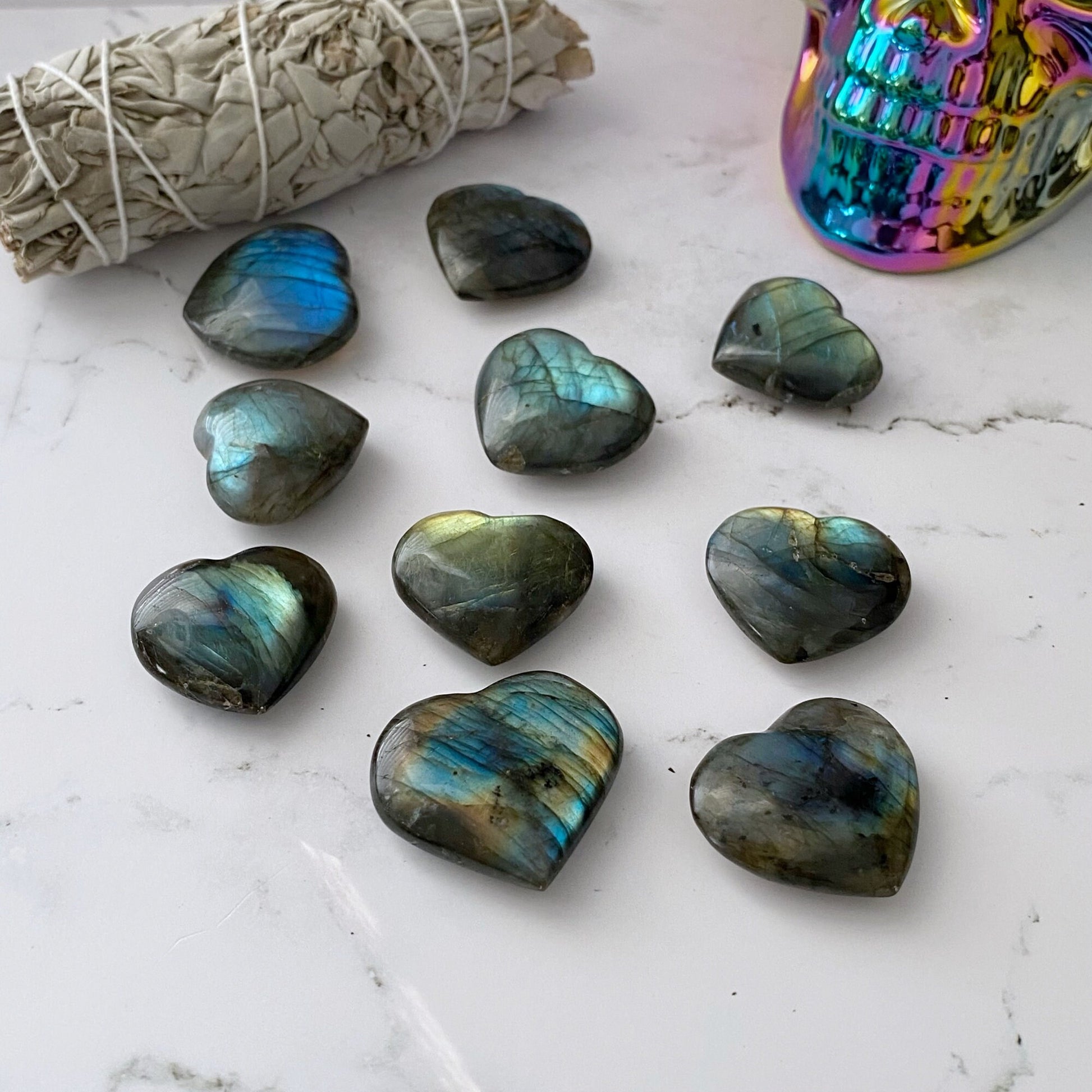 A collection of 1.5" Small Labradorite Hearts with Flash is arranged on a white marble surface. In the background, a bundle of sage and a multicolored, reflective skull decoration are visible.