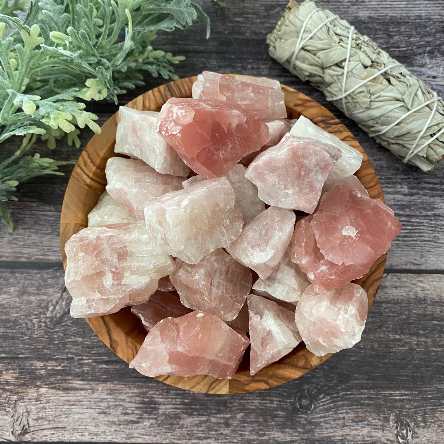 A wooden bowl filled with Raw Strawberry Pink Calcite Stones sits on a wooden surface. Next to the bowl is a bundle of dried sage and a sprig of green foliage, inviting a sense of inner peace and self-healing.