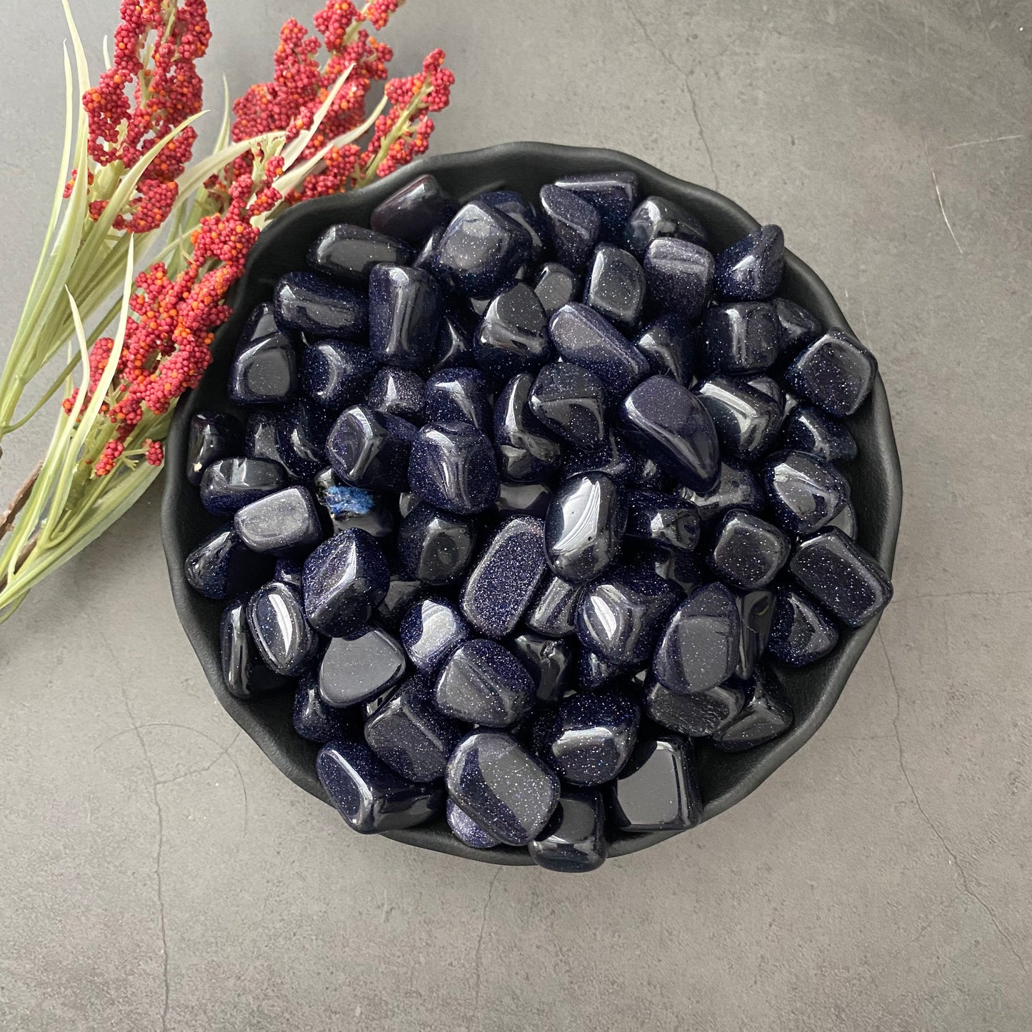 A black bowl filled with Blue Goldstone Tumbled Stones, known as a protective stone with a subtle sparkle, sits on a gray surface. To the left of the bowl, there is a sprig of red berries and green leaves. The overall scene is minimalistic and elegant.