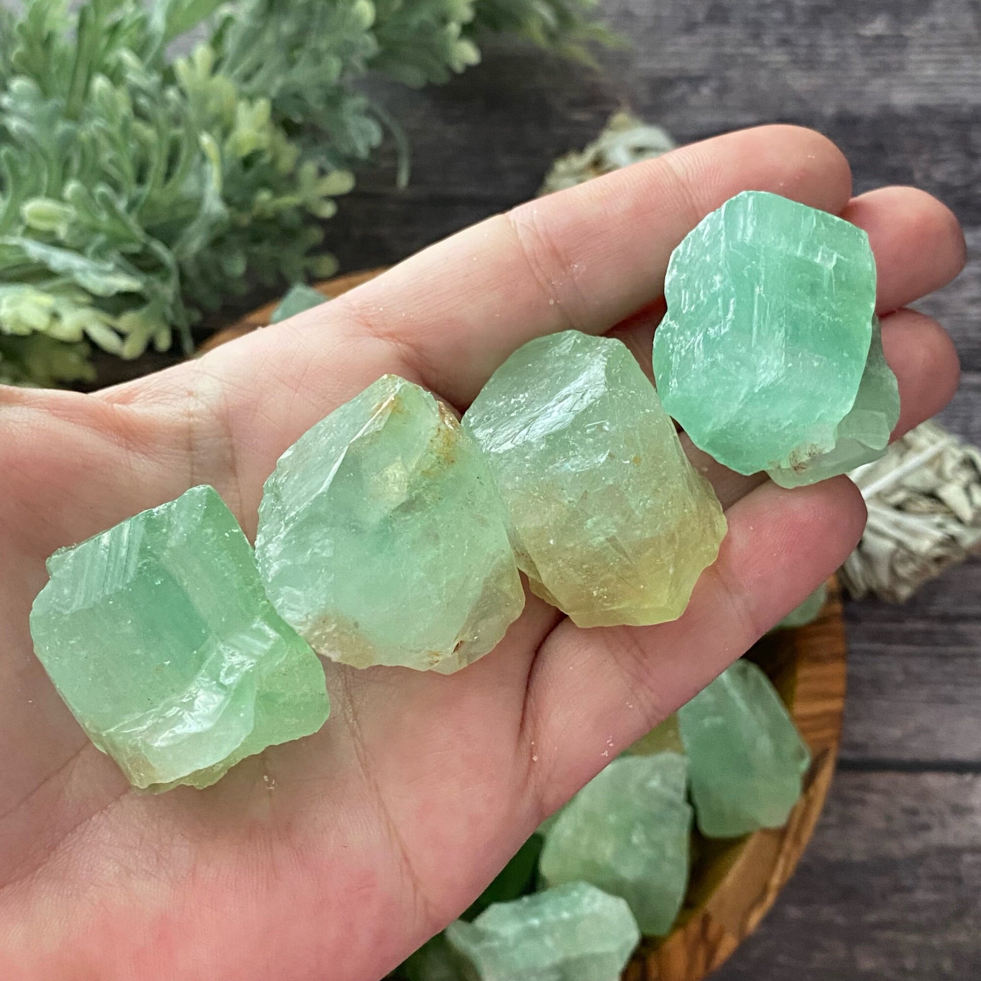 A wooden bowl filled with Raw Green Calcite Stones sits on a wooden surface. To the left of the bowl is some green foliage, and to the right, a bundle of sage wrapped in string. Known as a healing stone, Green Calcite symbolizes abundance and prosperity.