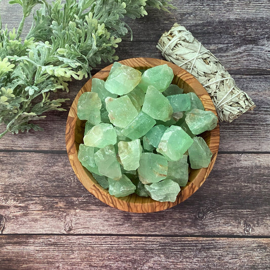 A wooden bowl filled with Raw Green Calcite Stones sits on a wooden surface. To the left of the bowl is some green foliage, and to the right, a bundle of sage wrapped in string. Known as a healing stone, Green Calcite symbolizes abundance and prosperity.