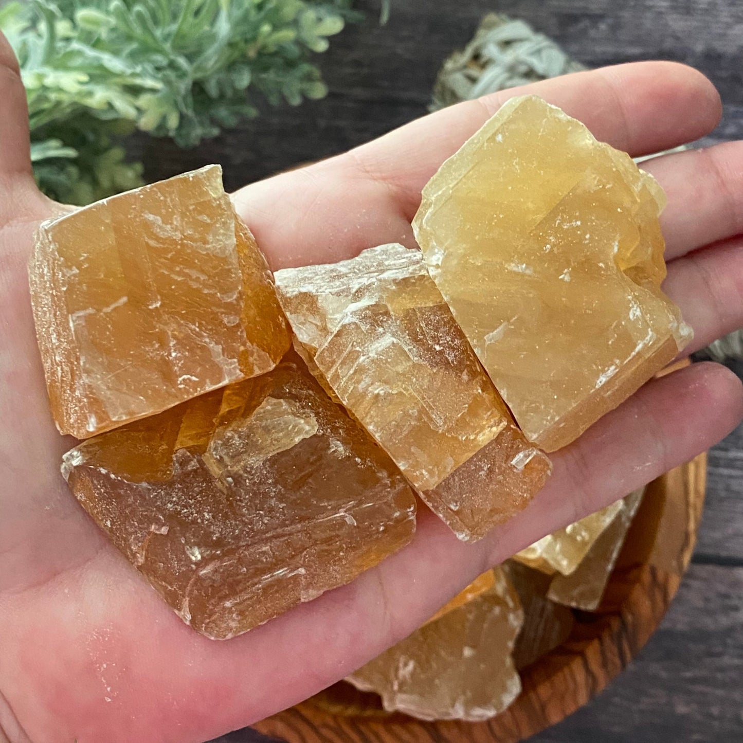 A wooden bowl filled with large, translucent Raw Honey Calcite Stones is placed on a rustic wooden surface. To the bowl’s right lies a smudge stick of bundled sage, while sprigs of green foliage decorate the left side, reinforcing an atmosphere of personal will and overcoming procrastination.