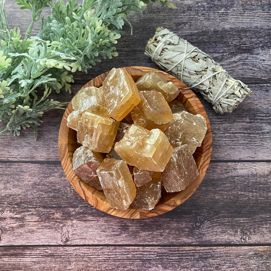 A wooden bowl filled with large, translucent Raw Honey Calcite Stones is placed on a rustic wooden surface. To the bowl’s right lies a smudge stick of bundled sage, while sprigs of green foliage decorate the left side, reinforcing an atmosphere of personal will and overcoming procrastination.