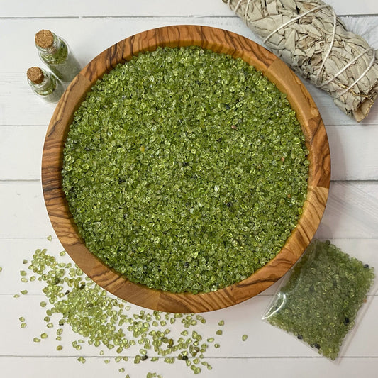 A wooden bowl filled with small green Peridot Crystal Chips, with some spilled on the white surface. Beside the bowl are two small vials containing similar crystals, a smudge stick for metaphysical uses, and a small plastic bag also containing the green crystals.